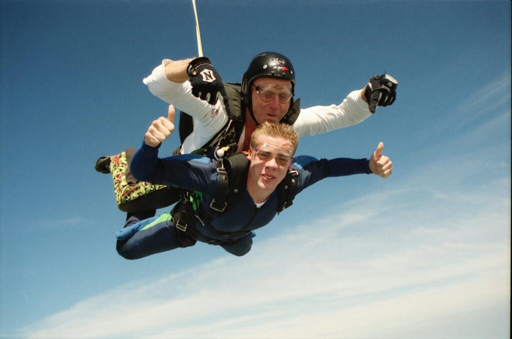 Queenstown, New Zealand. Two people tandem skydiving with thumbs up against a clear blue sky, embodying adventure and exhilaration.